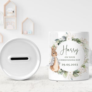 May include: White ceramic money box with a slot on the top. The front of the box features a watercolor illustration of a rabbit with a floral wreath and the text "Harry on your christening day 29.05.2023".