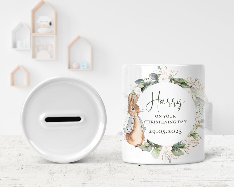 May include: White ceramic money box with a slot on the top. The front of the box features a watercolor illustration of a rabbit with a floral wreath and the text "Harry on your christening day 29.05.2023".