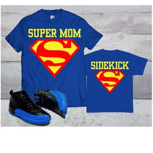 May include: Two blue t-shirts with a Superman logo. The larger shirt says "SUPER MOM" and the smaller shirt says "SIDEKICK". A pair of black and blue sneakers are in the bottom left corner.