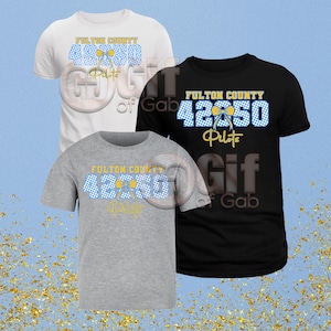 Puede incluir: Tres camisetas en blanco, gris y negro, cada una con el texto "FULTON COUNTY 42850 Pilots" en azul, amarillo y blanco. El diseño incluye un patrón de cuadros y un lazo.
