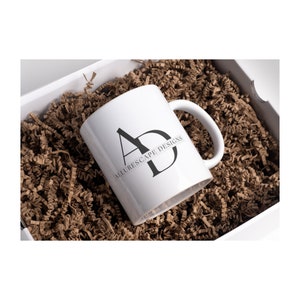 以下が含まれることがあります： 「Allurescape Designs」とスタイリッシュな「AD」のグラフィックが書かれた黒いロゴが付いた白いセラミックマグカップです。