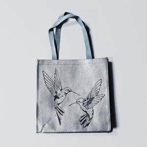 May include: A gray reusable tote bag with blue handles features a black and white illustration of two hummingbirds in flight.