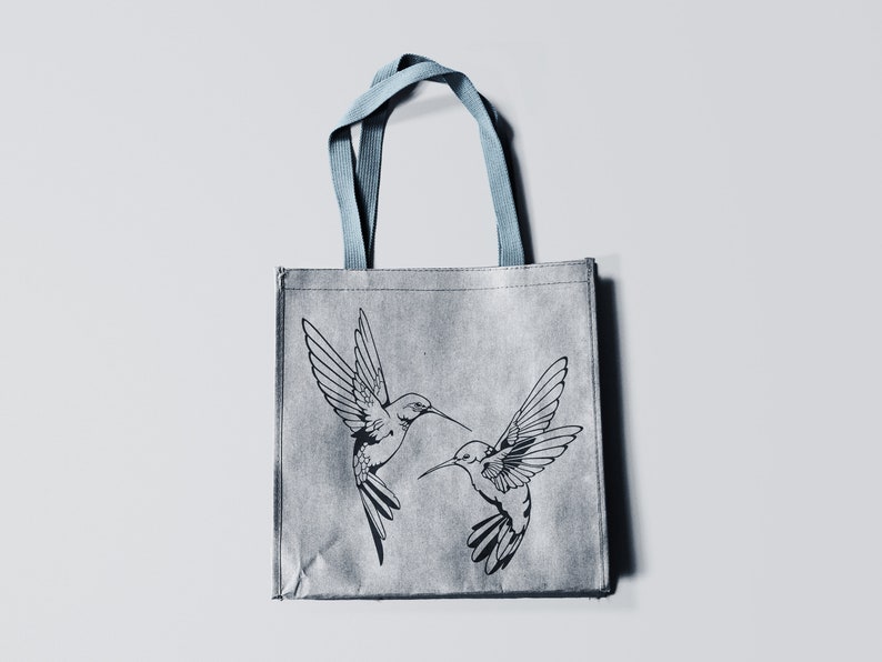 May include: A gray reusable tote bag with blue handles features a black and white illustration of two hummingbirds in flight.
