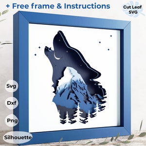 May include: A blue framed papercraft wall art featuring a wolf howling at the moon with a mountain range behind it. The wolf is silhouetted against a white background with stars and a crescent moon. The text "Free frame & Instructions" is at the top of the image.