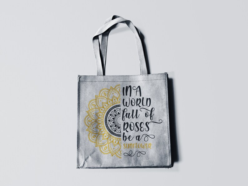 May include: A gray tote bag with a gold mandala design and the text "In a world full of roses be a sunflower" printed on it.