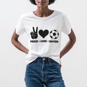 May include: White t-shirt with a black graphic design. The design includes a peace sign, a heart, and a soccer ball, with the words "PEACE LOVE SOCCER" below. The shirt is worn with blue jeans.