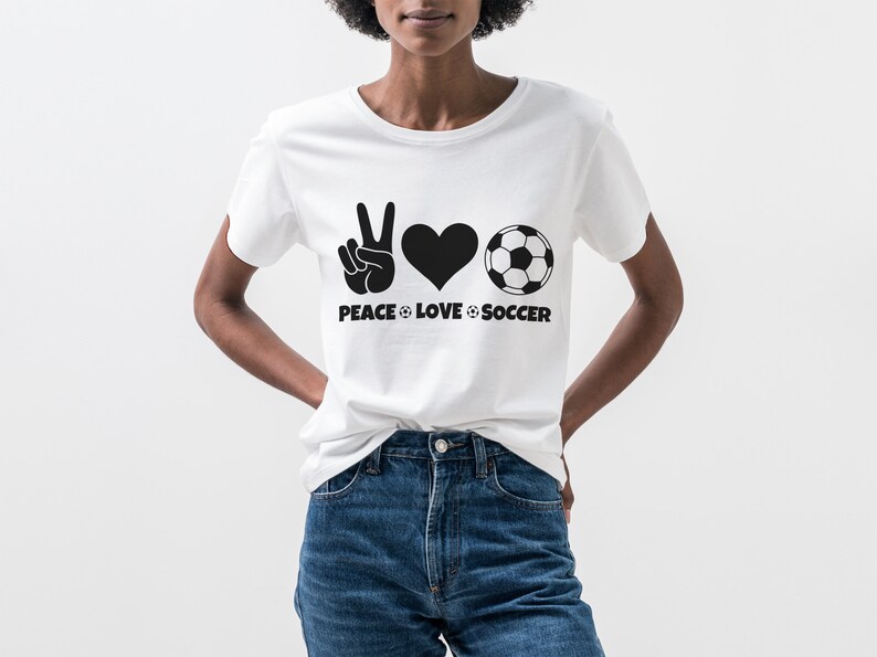 May include: White t-shirt with a black graphic design. The design includes a peace sign, a heart, and a soccer ball, with the words "PEACE LOVE SOCCER" below. The shirt is worn with blue jeans.