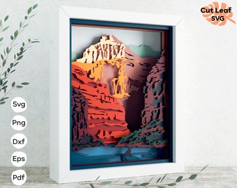 Puede incluir: Una obra de arte de papel recortado enmarcada que representa un paisaje en capas con un cañón, montañas y un río. La obra está hecha de papel de colores y tiene un efecto 3D.