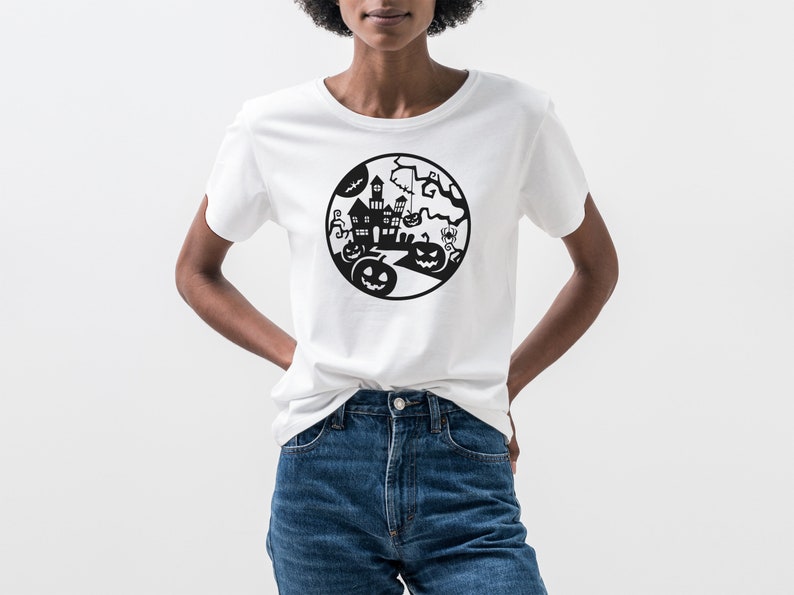 May include: White t-shirt featuring a black Halloween-themed graphic. The design includes a haunted house, pumpkins, bats, and a spider, all within a circular frame. The shirt is made of a soft material and is suitable for casual wear.