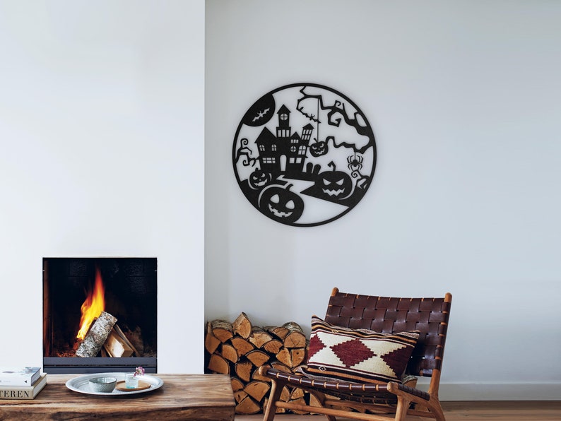 May include: Black metal Halloween wall art featuring a haunted house, pumpkins, a bat, and a spider within a circular frame. The artwork is displayed on a white wall above a wooden table and a leather chair.