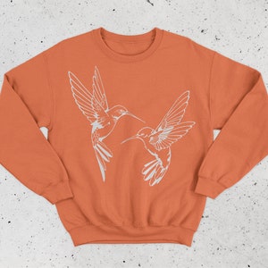 May include: A burnt orange sweatshirt with a white outline of two hummingbirds facing each other.