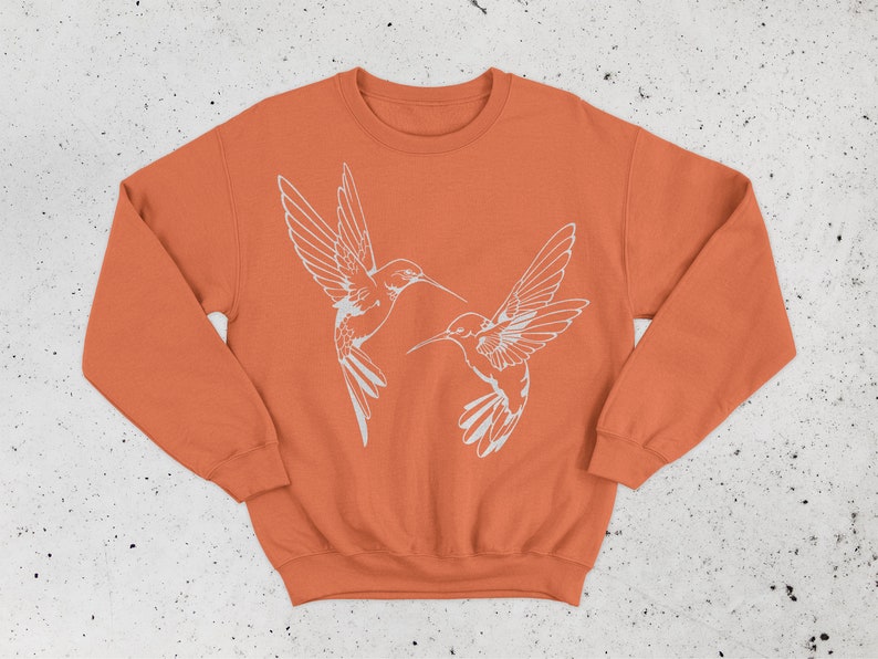 May include: A burnt orange sweatshirt with a white outline of two hummingbirds facing each other.