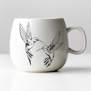 May include: White ceramic mug with a black line drawing of two hummingbirds in flight.