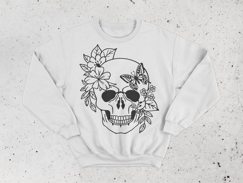 K&ouml;nnte beinhalten: Wei&szlig;er Pullover mit einem schwarz-wei&szlig;en Motiv eines Sch&auml;dels, der mit Blumen und einem Schmetterling verziert ist.