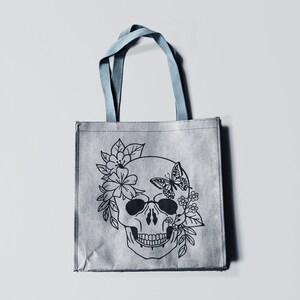 K&ouml;nnte beinhalten: Eine graue Einkaufstasche mit einer schwarz-wei&szlig;en Illustration eines Sch&auml;dels mit Blumen und einem Schmetterling auf der Vorderseite.