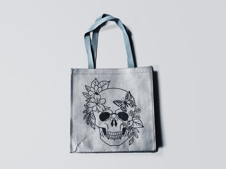 K&ouml;nnte beinhalten: Eine graue Einkaufstasche mit einer schwarz-wei&szlig;en Illustration eines Sch&auml;dels mit Blumen und einem Schmetterling auf der Vorderseite.