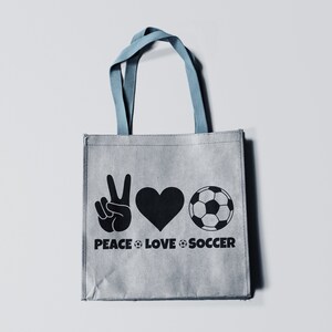 May include: Gray tote bag with blue handles featuring a black graphic of a peace sign, heart, and soccer ball. The words "PEACE LOVE SOCCER" are printed below the symbols. The bag is made of a textured material.