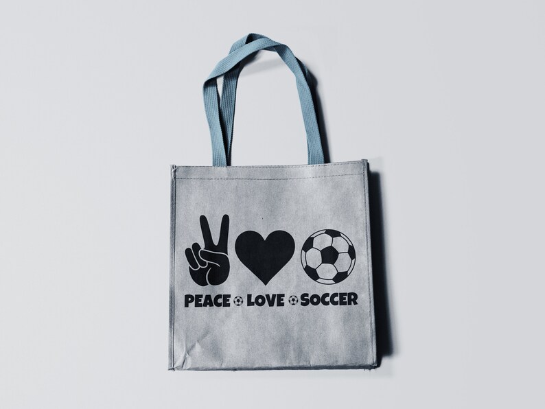 May include: Gray tote bag with blue handles featuring a black graphic of a peace sign, heart, and soccer ball. The words "PEACE LOVE SOCCER" are printed below the symbols. The bag is made of a textured material.