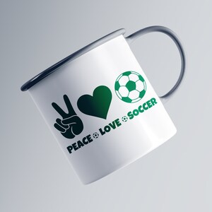 May include: White enamel mug with a dark gray handle and rim. The mug features a green peace sign, heart, and soccer ball graphic with the words "PEACE LOVE SOCCER" in green. The mug is angled slightly to the left.