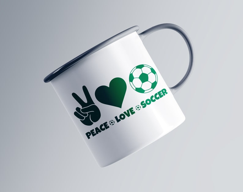 May include: White enamel mug with a dark gray handle and rim. The mug features a green peace sign, heart, and soccer ball graphic with the words "PEACE LOVE SOCCER" in green. The mug is angled slightly to the left.