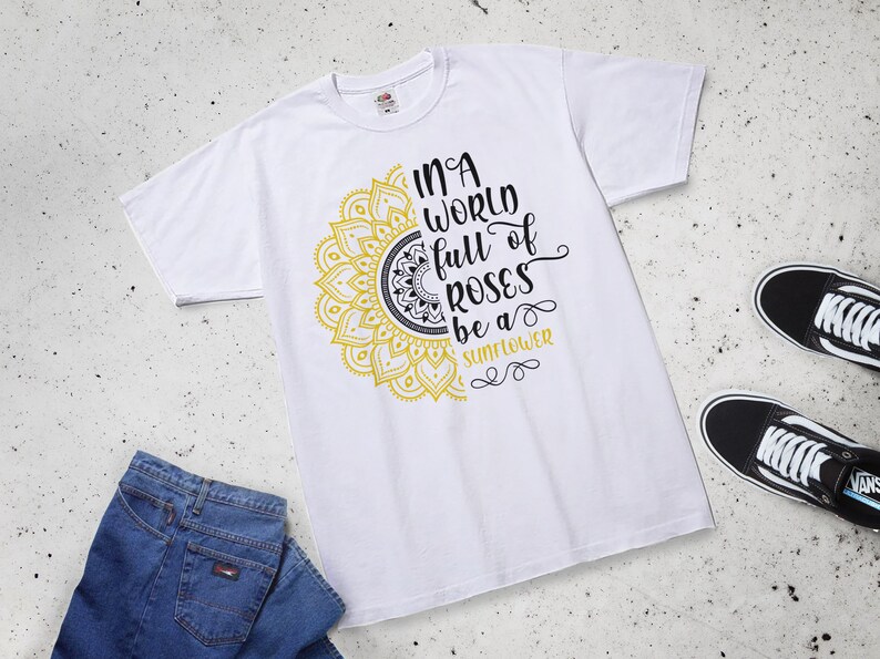 May include: White t-shirt with a yellow and black mandala design and the text "In a world full of roses be a sunflower".