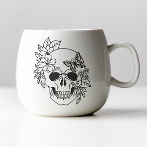 K&ouml;nnte beinhalten: Wei&szlig;e Keramiktasse mit einer schwarzen Linienzeichnung eines Sch&auml;dels mit Blumen und einem Schmetterling auf einer Seite.