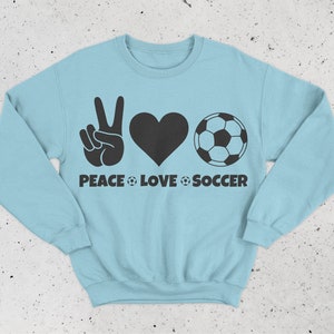 May include: Light blue sweatshirt with a black graphic design. The design includes a peace sign, a heart, and a soccer ball, with the words "PEACE LOVE SOCCER" below. The sweatshirt is a crew neck style.