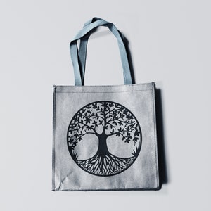 May include: A gray reusable tote bag with blue handles features a black tree of life design. The tree is stylized with a thick trunk and intricate roots.