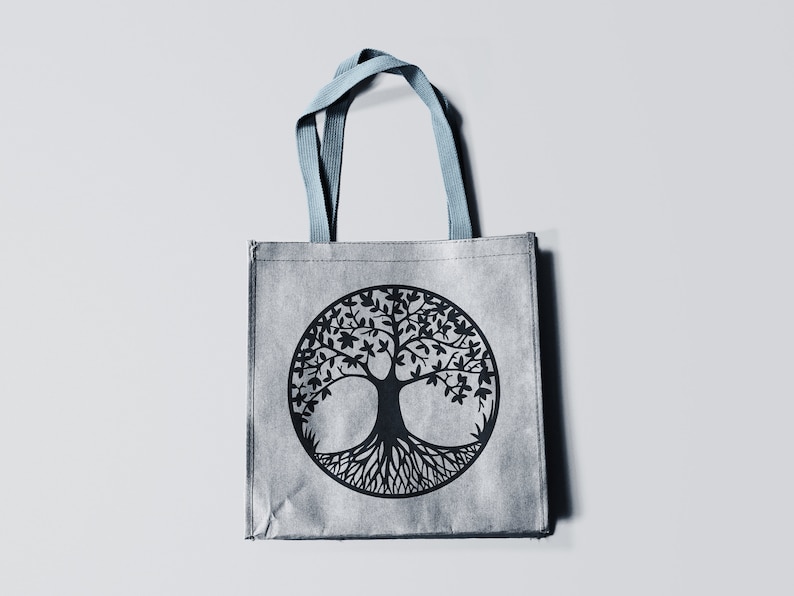 May include: A gray reusable tote bag with blue handles features a black tree of life design. The tree is stylized with a thick trunk and intricate roots.