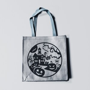 May include: Gray Halloween tote bag with a black circular design featuring a haunted house, pumpkins, bats, and a spider. The bag has blue handles and is made of a textured material.