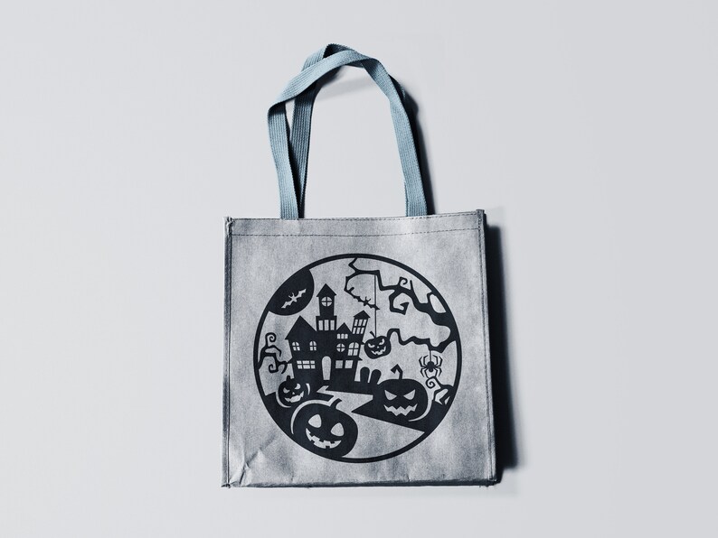 May include: Gray Halloween tote bag with a black circular design featuring a haunted house, pumpkins, bats, and a spider. The bag has blue handles and is made of a textured material.
