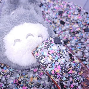 Puede incluir: Primer plano de un juguete de peluche gris con una cara sonriente, parcialmente cubierto por una funda de tel&eacute;fono transparente llena de abalorios de resina de colores y negros. Los abalorios incluyen estrellas, animales peque&ntilde;os y n&uacute;meros.