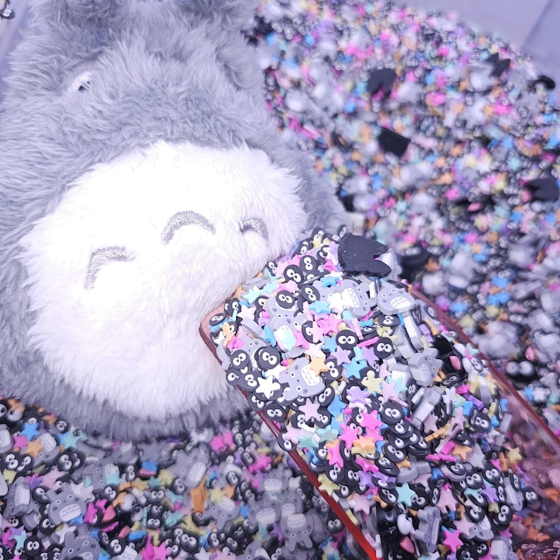 Puede incluir: Primer plano de un juguete de peluche gris con una cara sonriente, parcialmente cubierto por una funda de tel&eacute;fono transparente llena de abalorios de resina de colores y negros. Los abalorios incluyen estrellas, animales peque&ntilde;os y n&uacute;meros.