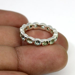 May include: A silver ring with a continuous row of oval-cut, light green gemstones. The ring is set in a polished silver band, creating a delicate design. The gemstones are evenly spaced around the ring's circumference.