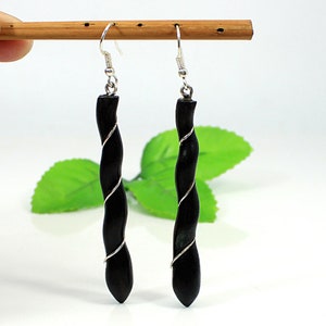 May include: A pair of black wooden earrings with a twisted design, wrapped with silver-coloured wire. The earrings hang from silver-coloured hooks. The background is white with a green leaf.