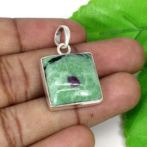 Puede incluir: Colgante cuadrado con una piedra verde, inclusiones negras y rojas, engastado en un marco de metal plateado. El colgante tiene un lazo en la parte superior para una cadena. La superficie de la piedra está pulida y tiene un aspecto natural y texturizado.