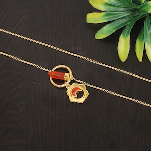 May include: A gold chain necklace with a pendant featuring a red carnelian stone in a circle and a crescent moon design.