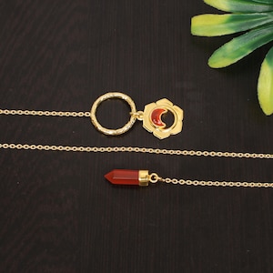May include: A gold chain necklace with two pendants. One pendant is a circle with a textured surface and the other is a flower-shaped design with a red crescent moon in the center.