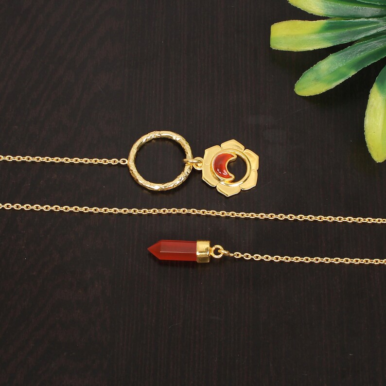 May include: A gold chain necklace with two pendants. One pendant is a circle with a textured surface and the other is a flower-shaped design with a red crescent moon in the center.