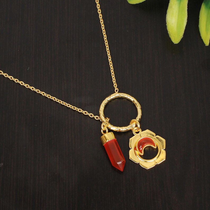 May include: A gold necklace with a red gemstone pendant and a gold charm with a crescent moon design. The charm is attached to a gold ring.
