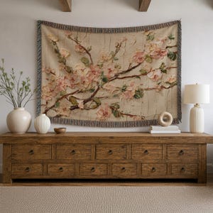 May include: A woven wall tapestry featuring a textured floral design with light pink and cream blossoms on dark brown branches. The rectangular textile has a fringed edge with multi-colored threads and is displayed horizontally on a wall.