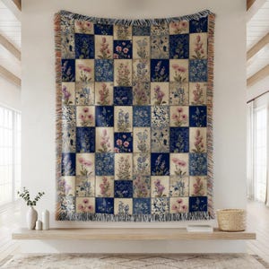May include: A woven throw blanket featuring a patchwork grid design with alternating squares of cream and deep blue. Each square displays detailed botanical illustrations of various flowers in shades of pink, purple, and blue, finished with fringe.