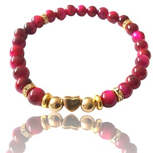 Könnte beinhalten: Ein rosafarbenes Tigerauge-Armband mit einem goldenen Herz-Anhänger. Das Armband besteht aus runden Perlen mit glänzender Oberfläche.