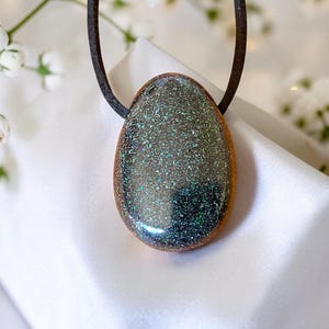 Könnte beinhalten: Ein tropfenförmiger Anhänger mit einer dunklen, glitzernden Oberfläche mit irisierenden grünen und blauen Sprenkeln, eingefasst in einen glatten, polierten Holzrahmen. Der Anhänger hängt an einer schlichten, dunkelbraunen Kordelkette.
