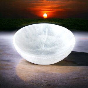 May include: A white selenite bowl, also known as a sound bowl, is shown on a light brown surface. The bowl is smooth and has a slight sheen. The background is blurred and shows a sunset with a bright orange sky.