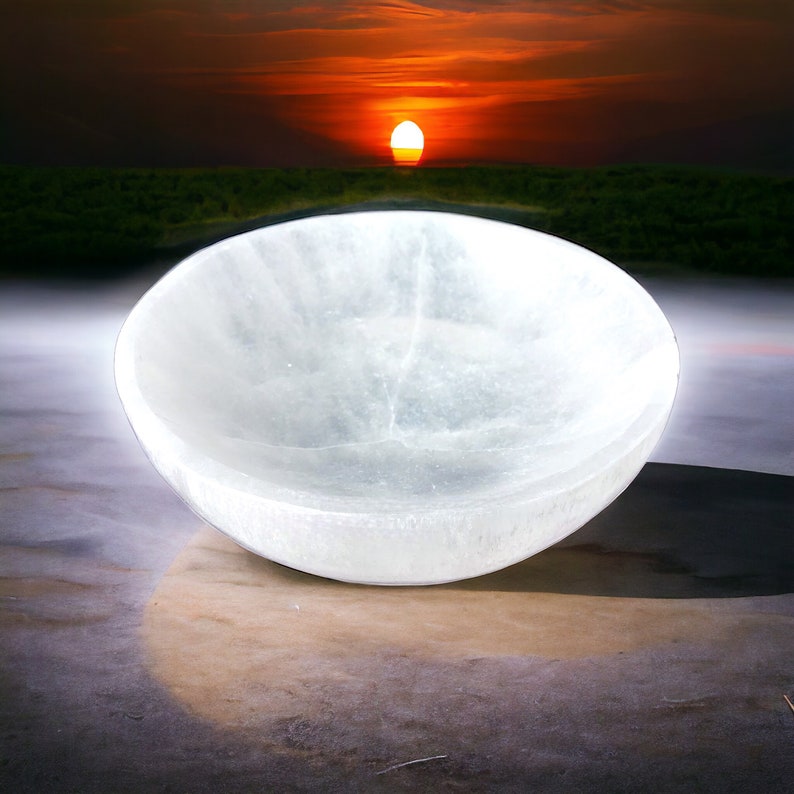 May include: A white selenite bowl, also known as a sound bowl, is shown on a light brown surface. The bowl is smooth and has a slight sheen. The background is blurred and shows a sunset with a bright orange sky.