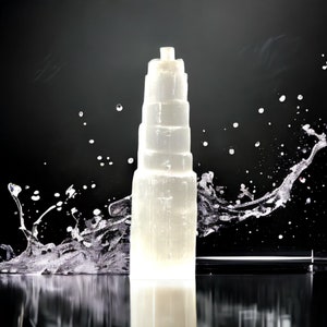 Könnte beinhalten: Ein weißer Selenit-Turm mit einem geschichteten, abgestuften Design. Der Turm steht auf einer schwarzen Oberfläche, um die herum Wasser spritzt.