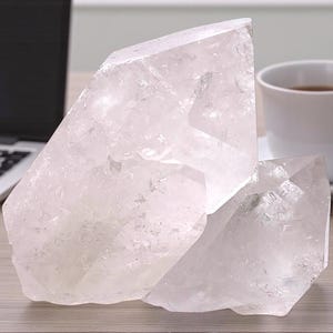 Könnte beinhalten: Zwei große, klare Quarz-Kristallcluster liegen auf einer hellen Holzoberfläche. Eine weiße Tasse und Untertasse mit dunkler Flüssigkeit und ein Laptop befinden sich im Hintergrund. Die Kristalle haben eine raue, natürliche Textur.