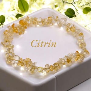 Könnte beinhalten: Ein Citrin-Armband aus kleinen, unregelmäßigen Citrin-Stücken. Das Armband wird auf einer weißen Oberfläche mit dem Wort "Citrin" in goldenen Buchstaben präsentiert.