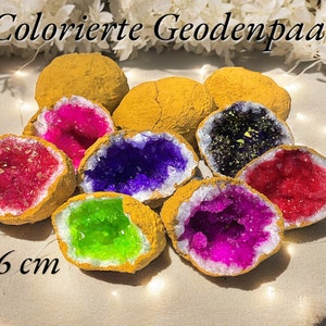 Può includere: Otto metà di geodi, ciascuna con un cristallo di colore diverso all'interno. I geodi hanno un diametro di circa 5-6 cm e sono dipinti di un colore giallo dorato. "Colorierte Geodenpaare" è scritto in tedesco nella parte superiore dell'immagine.